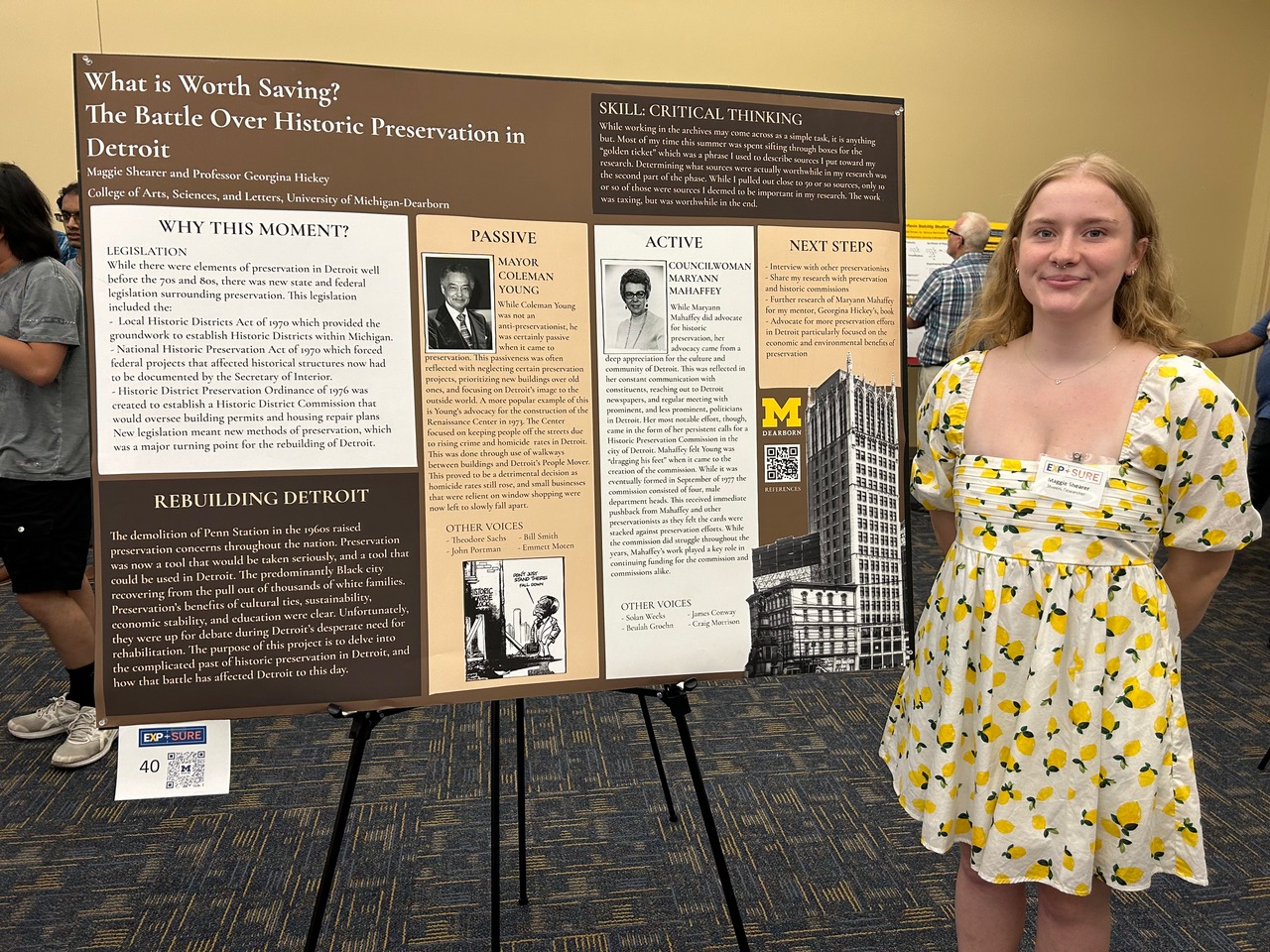 a student with long light hair stands beside a research poster presentation, wearing a white dress with yellow lemons on it