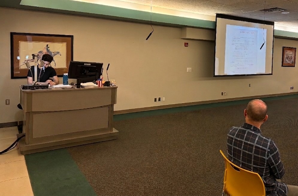 A presenter sits at a desk with a computer and microphone in a conference room.A large projection screen displays a document with lines of text.An attendee sits in the foreground facing the presenter and screen.