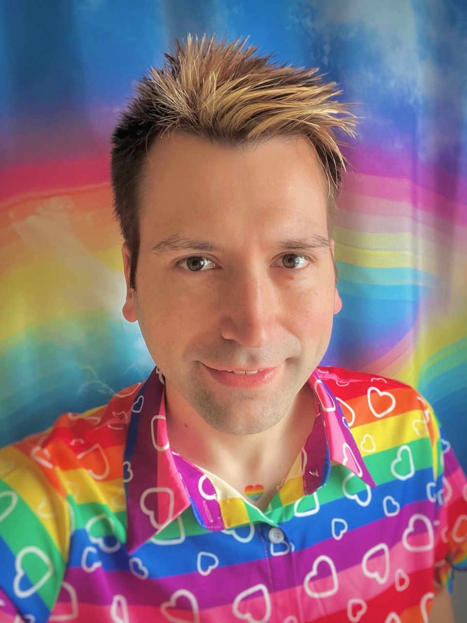 a person with blonde and brown spiked hair smiles in front of a rainbow background wearing  rainbow button-up shirt with hearts on it