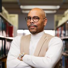 Individual standing in front of books in a white long sleeve shirt and beige vest.