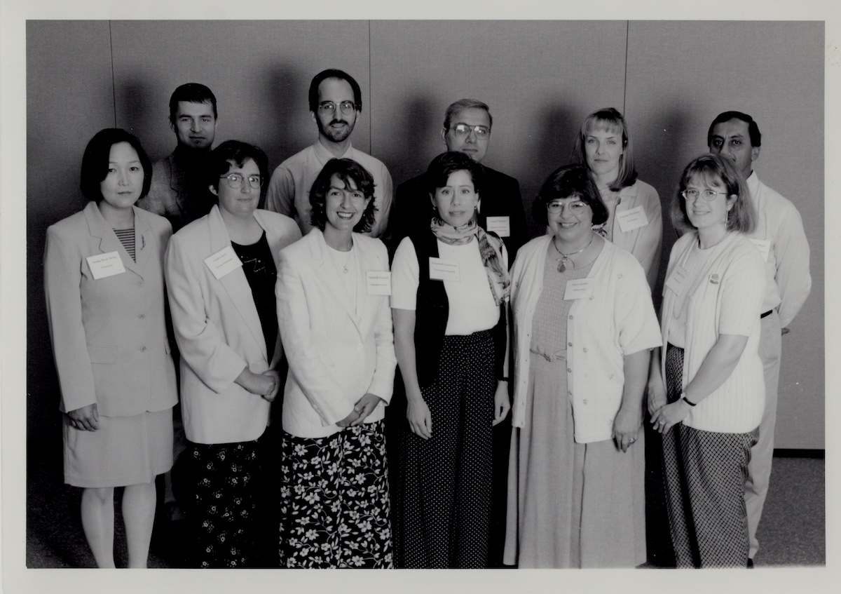 A black-and-white portrait of ten people arranged in two rows against a neutral wall, dressed in late 1990s business-casual attire with name tags pinned to their clothes. The front row consists of five women smiling, while the back row features four men and one woman standing behind them.