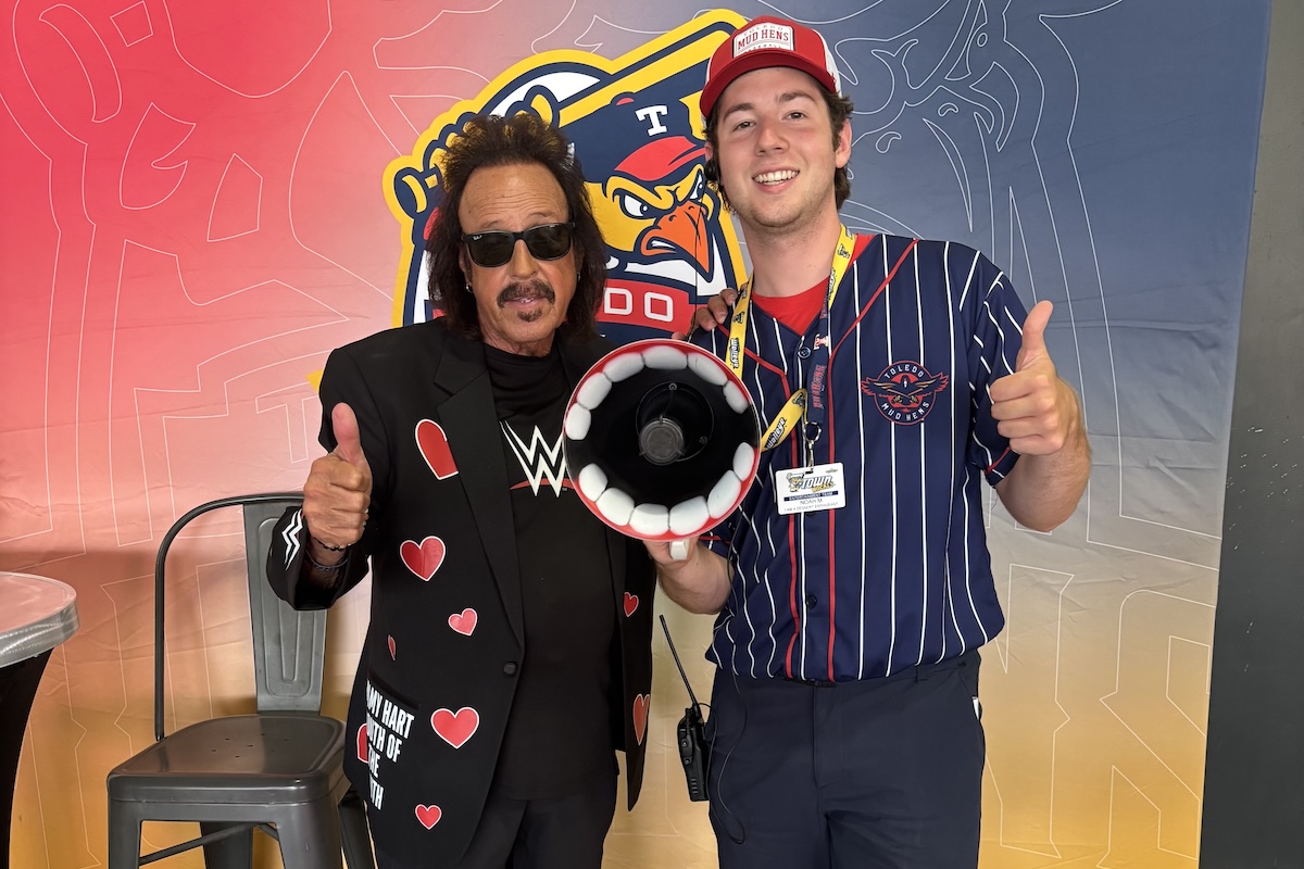 A smiling young man in a navy Toledo Mud Hens baseball jersey, hat, and staff lanyard poses next to WWE legend Jimmy "The Mouth of the South" Hart. Both give a thumbs-up gesture while the young man holds Hart's iconic red-and-white megaphone between them in front of a Toledo Mud Hens backdrop.