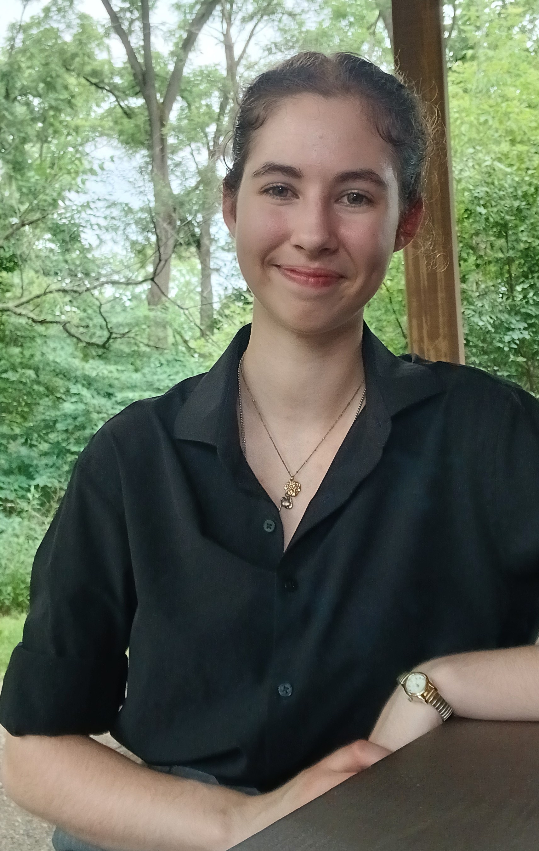 Person in a black button-up shirt sits at an outdoor wooden table.They wear a gold necklace and a wristwatch, with arms resting on the table.Leafy green trees and a wooden post fill the background.