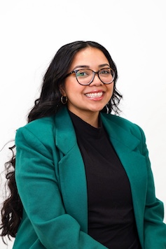 a smiling person with long dark hair and glasses pictured in front a white backdrop wearing a teal blazer and black top