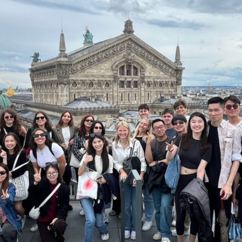 A group of students poses on a rooftop terrace in Paris with the Palais Garnier (Opera House) in the background. Everyone’s smiling and it looks like a fun study trip moment.