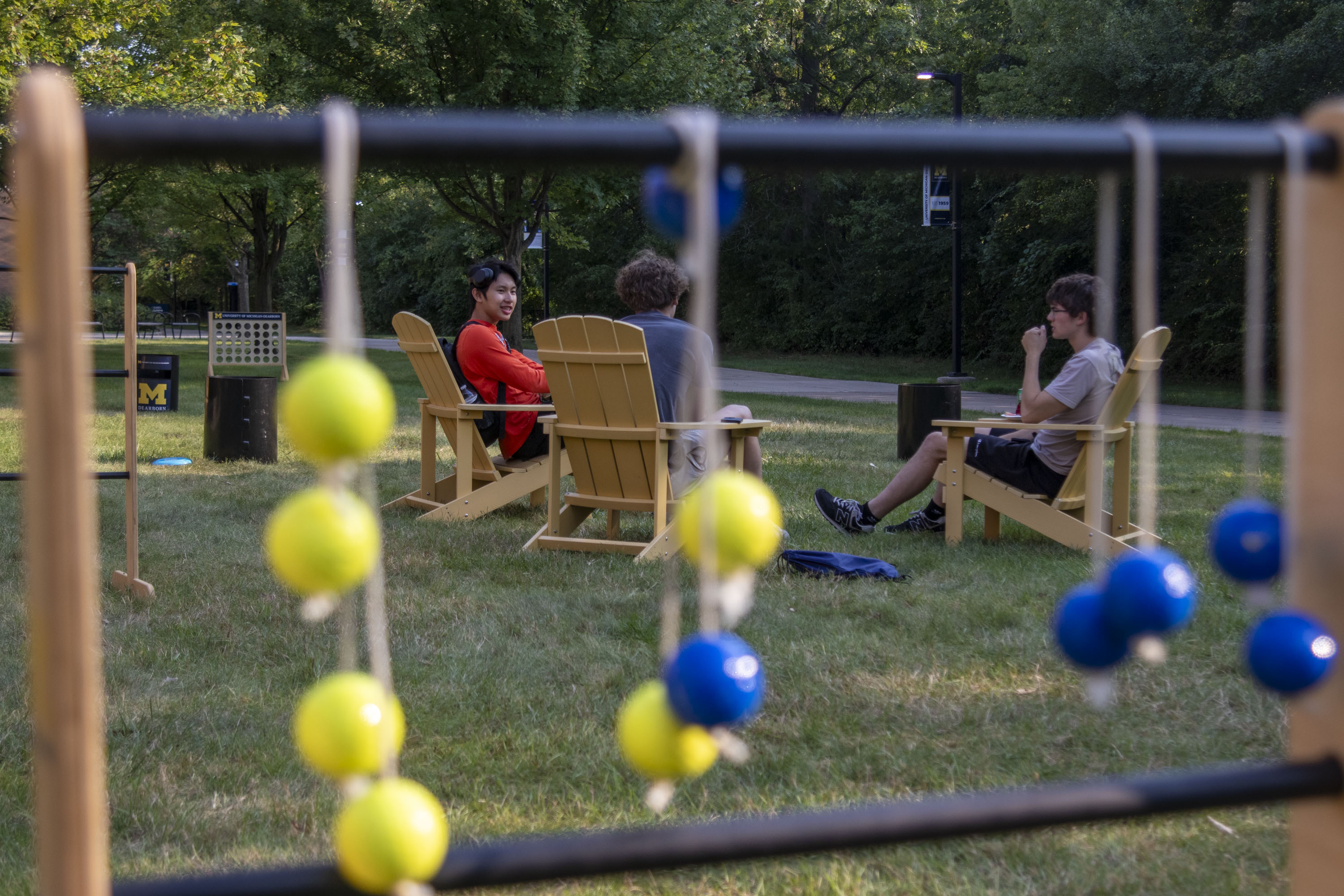 Students sit in wooden Adirondack chairs in the shade, with lawn games in the foreground and background