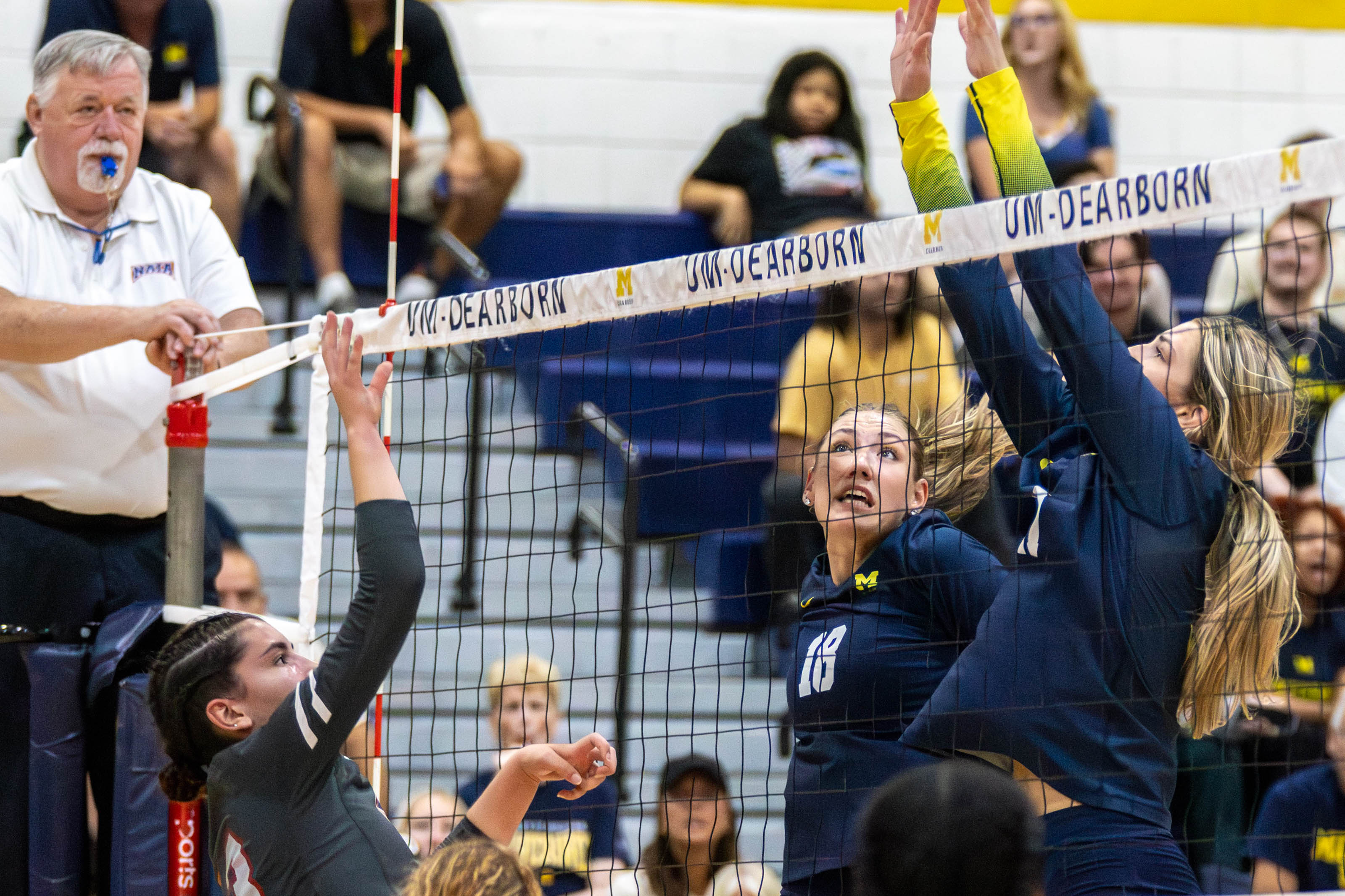 A women's volleyball player jumps to spike a ball with referee with a whistle in his mouth in the background