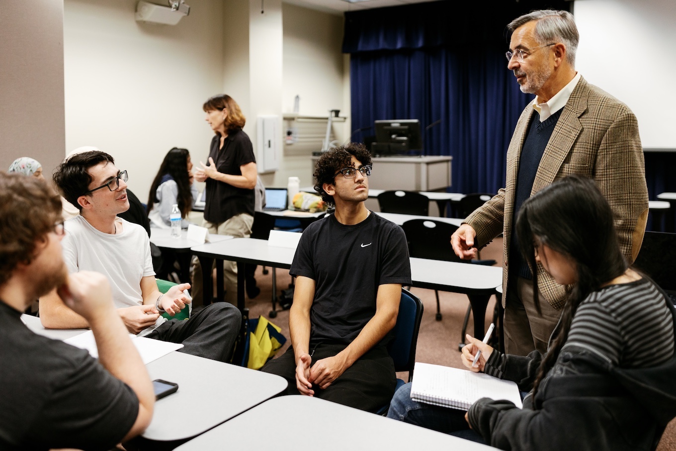 A group of seated students speak to a male professor in a beige suit in a classroom.