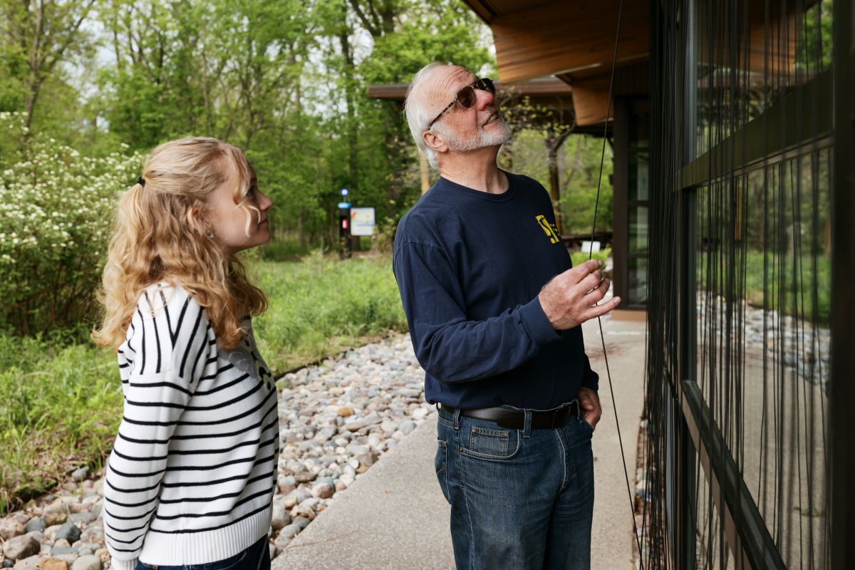 Two people stand outside a building examining vertical paracord lines installed in front of reflective windows to help prevent bird-window collisions.