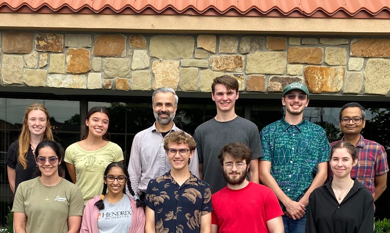 A group of people pose together outdoors in front of a stone wall.Casual attire and varied expressions suggest an informal gathering or group photo.Greenery and reflections are visible through the window behind the group.