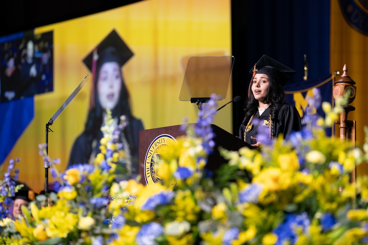 A woman stands at a podium and speaks. She is wearing a UM-Dearborn graduation cap and gown. Behind her, there is a large projection screen with her photo.