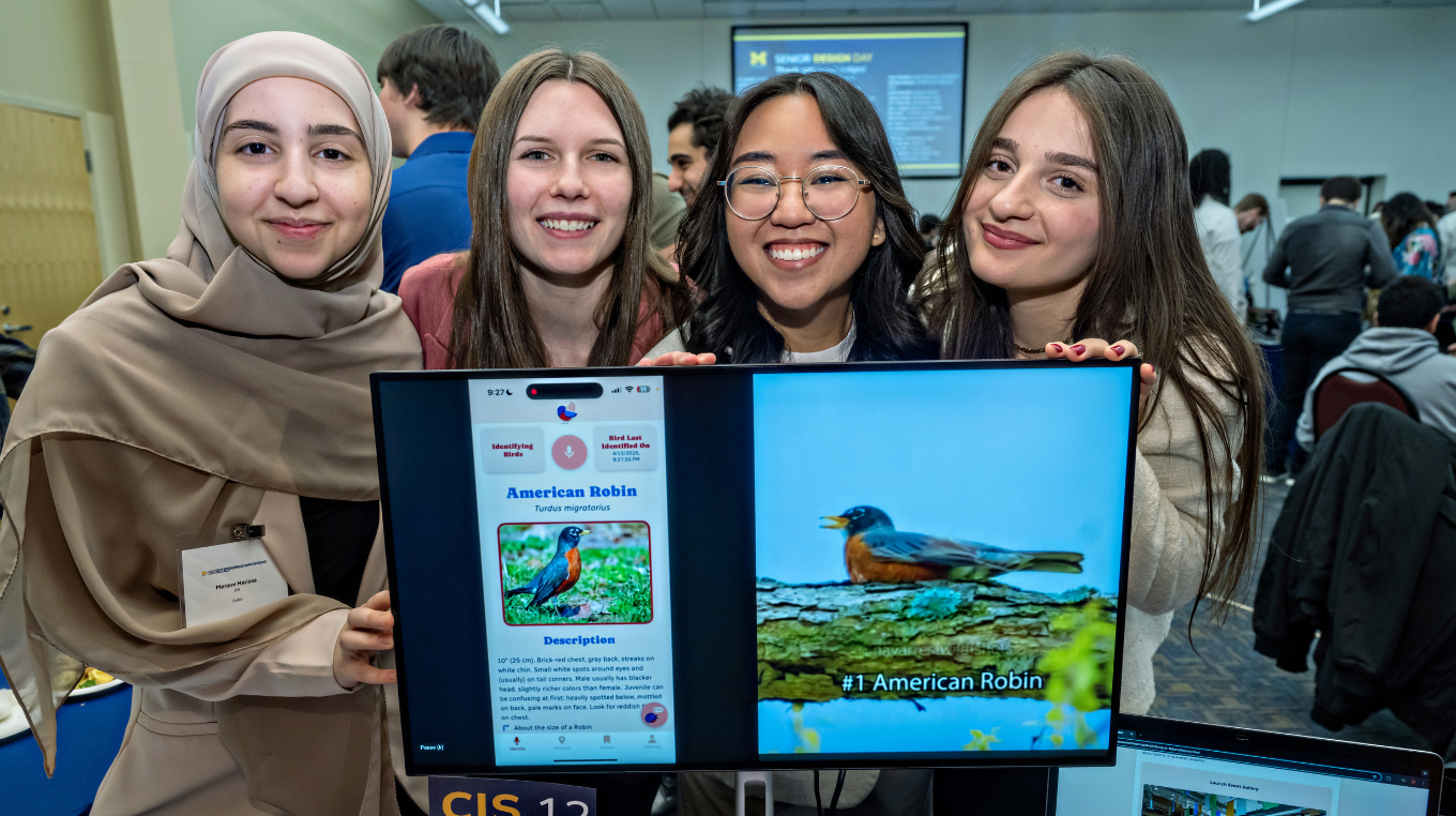 Four smiling students pose behind a monitor displaying a mobile bird-identification app and a photo of an American robin at a crowded showcase.