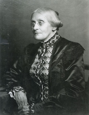 lack and white portrait of an elderly woman seated, wearing a dark satin dress with lace trim and a high-necked lace blouse. Her hair is styled in a low bun and she rests one hand on her lap.