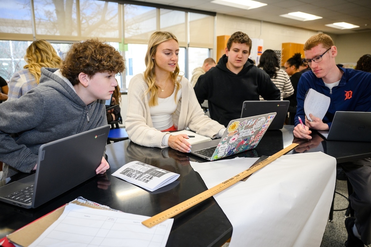 Four people gather around a table to look at a laptop