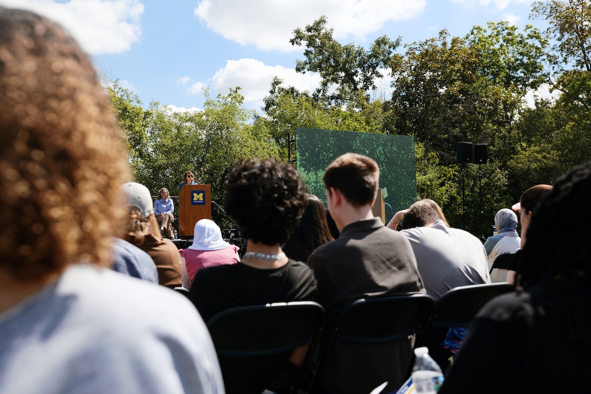An audience seated outdoors listening to a speaker at a podium featuring a UM-Dearborn logo.