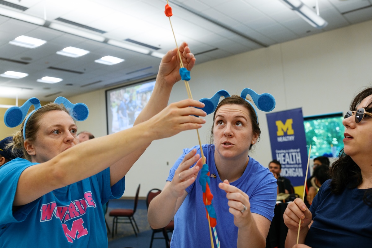 People wearing blue antenna headbands collaborate indoors on constructing a tall structure out of wooden skewers and modeling clay.