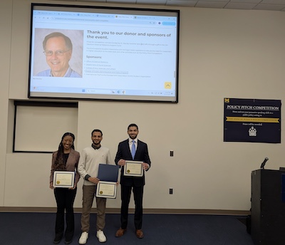3 smiling people stand holding certificates in front of a display screen and a poster about the Policy Pitch Competition