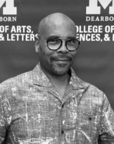 A black-and-white portrait of a man smiling at the camera, wearing glasses and a patterned button-up shirt. He stands in front of a UM-Dearborn College of Arts, Sciences, & Letters backdrop.