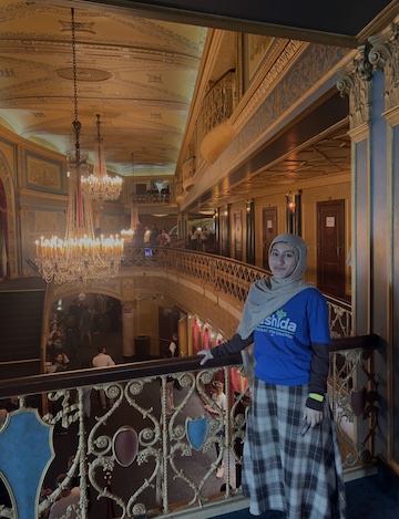 A student participating in the Rep. Rashida Tlaib Democracy Summer Fellowship stands on an ornate balcony inside a historic theater while wearing a blue fellowship T-shirt. Decorative architecture, chandeliers and a view of the theater below provide the backdrop