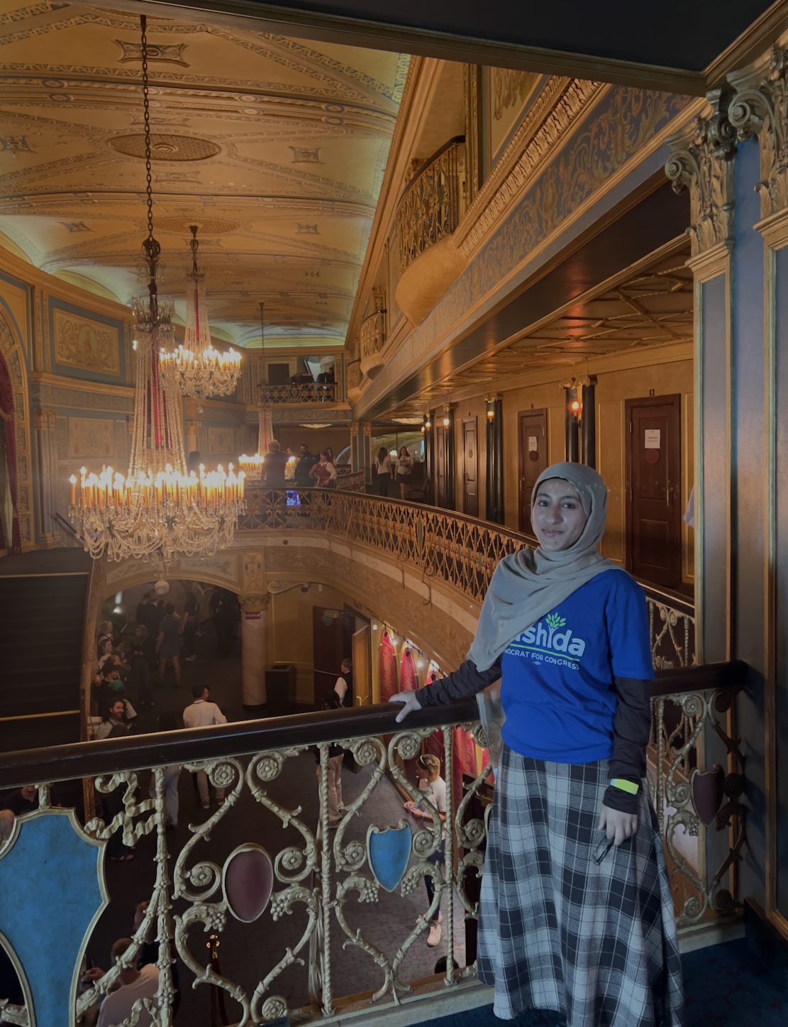 A student participating in the Rep. Rashida Tlaib Democracy Summer Fellowship stands on an ornate balcony inside a historic theater while wearing a blue fellowship T-shirt. Decorative architecture, chandeliers and a view of the theater below provide the backdrop
