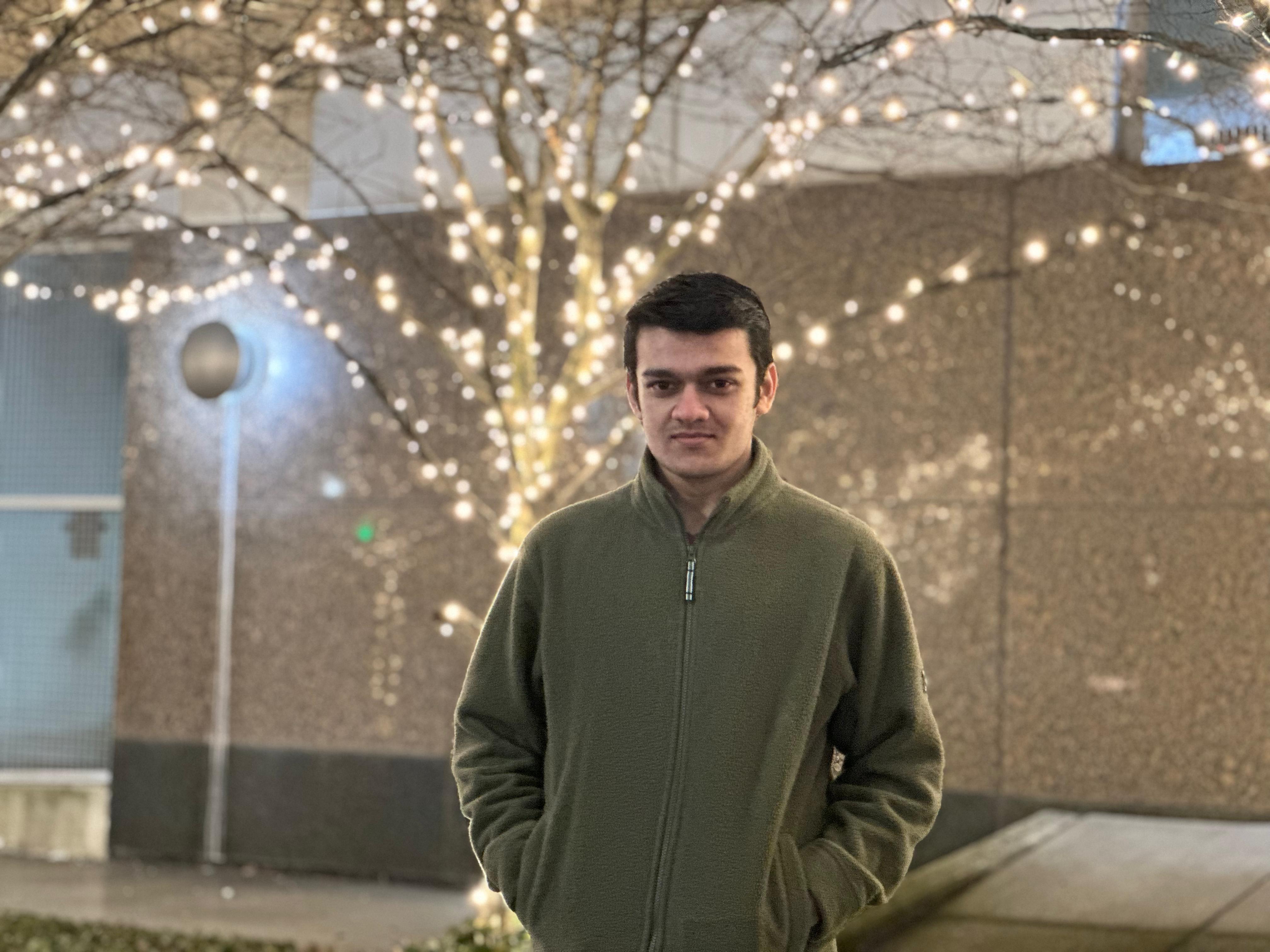 Shubh Bhojak standing outside in a green sweater. Lights are on the trees behind him and a building behind the tree.