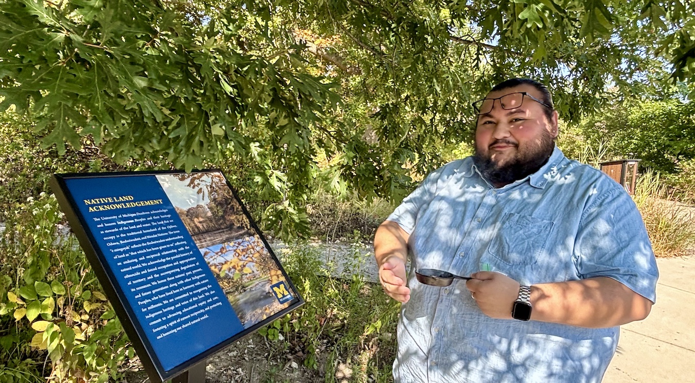 A man holding a small pan burns white sage in an outdoor area of campus.
