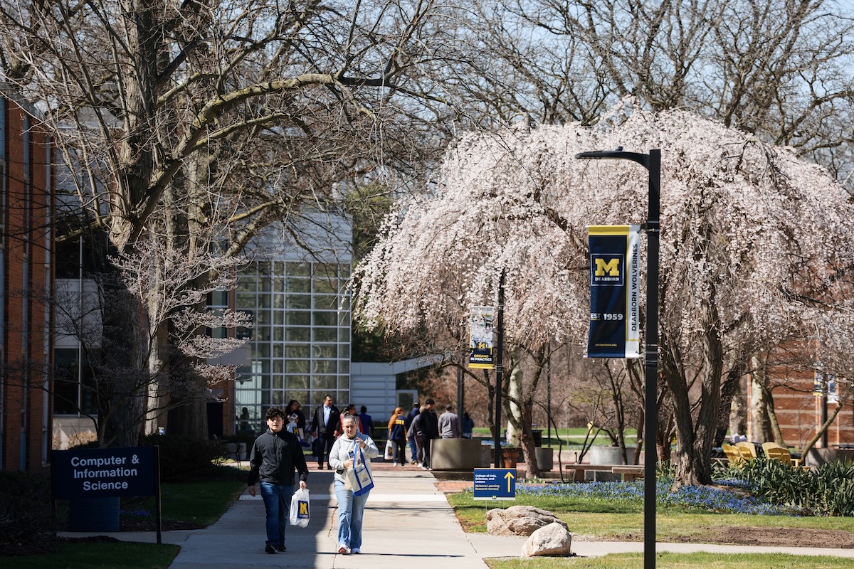 A photo of the UM-Dearborn campus that includes white flowering trees, the exterior of a building that has floor to ceiling windows and many students walking on campus walkways 
