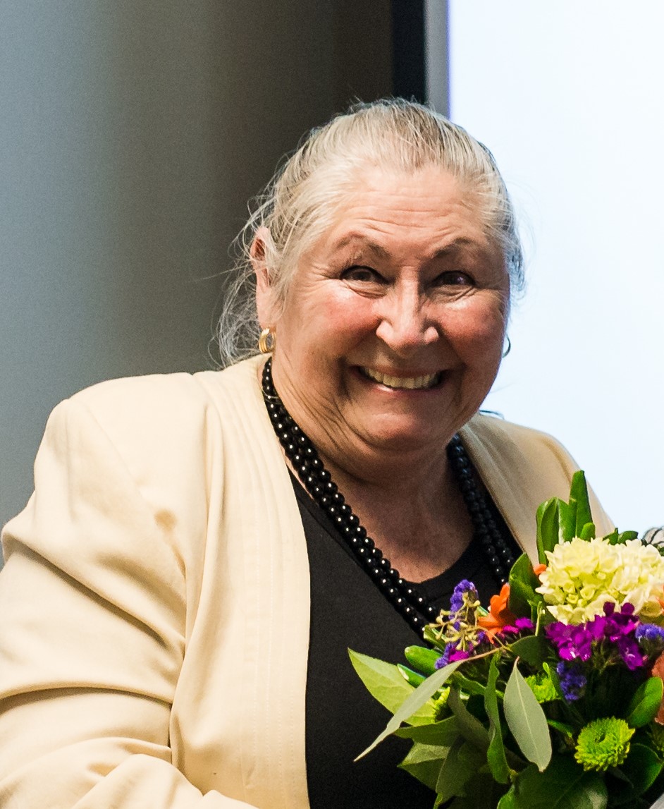 A person wearing a cream-colored blazer and black top holds a vibrant bouquet of flowers with purple, orange, and green blooms. Gold earrings and a black beaded necklace complete the outfit, shown against a softly lit indoor background.