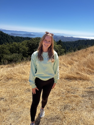woman with brownish hair stands in front of mountains and lakes on dry straw wearing black pants and a mint green shirt