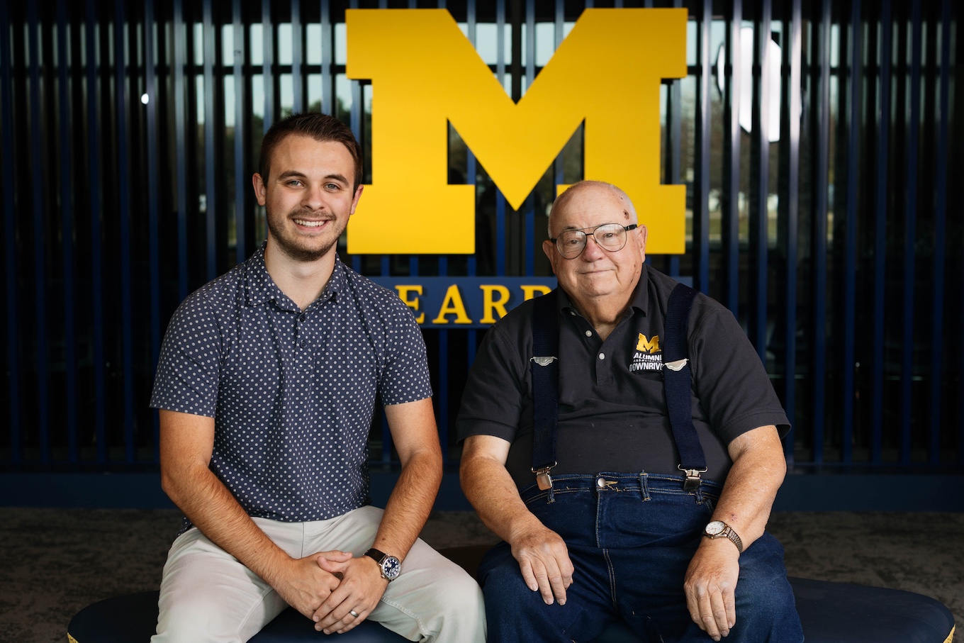 Two men sit together with a UM-Dearborn block M in the background.