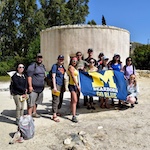 A group of students standing outdoors at an archaeological ruin with a large circular stone structure, holding a University of Michigan–Dearborn banner on a sunny day.