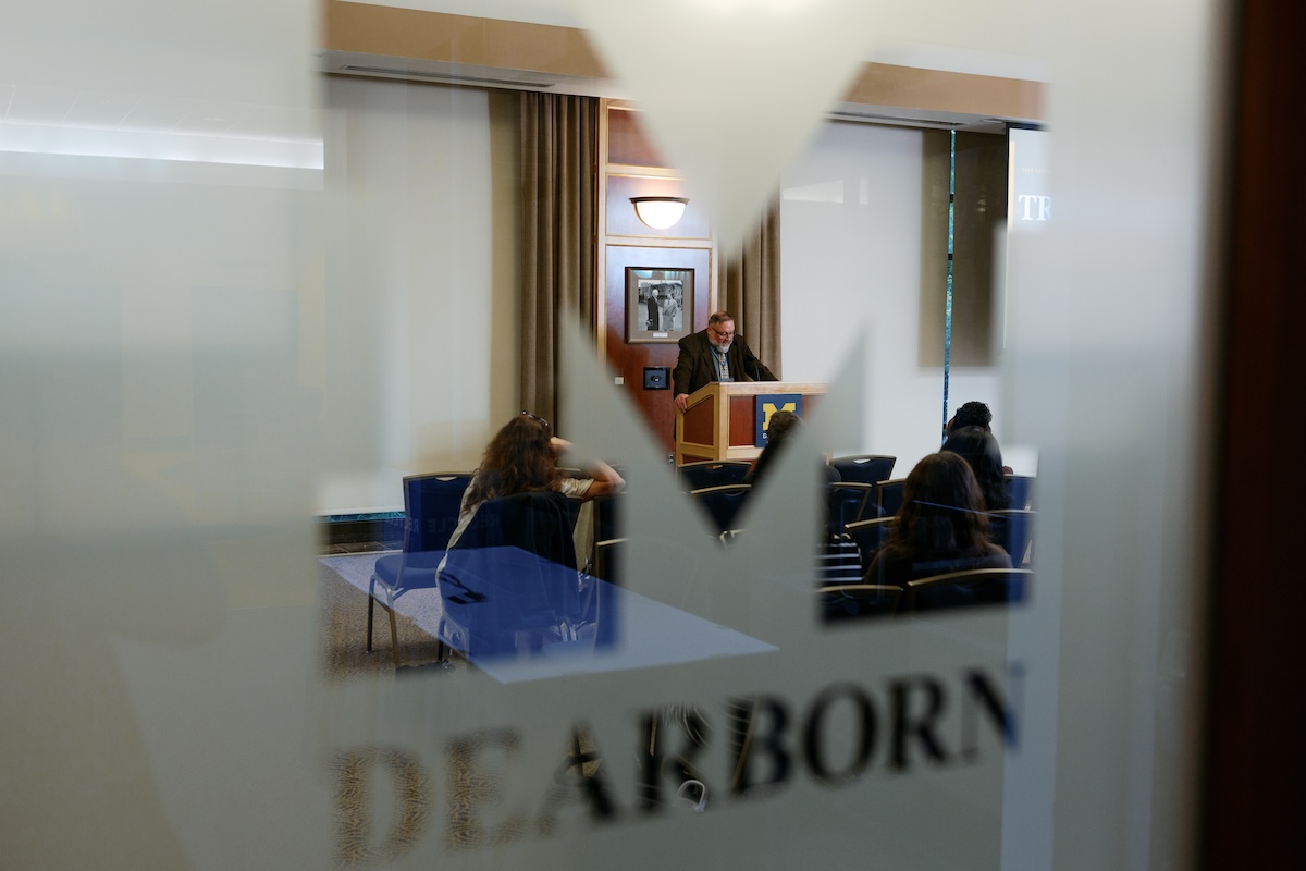 A speaker addresses an audience at a podium, viewed through a frosted glass window featuring the "Block M" logo and the word "DEARBORN."