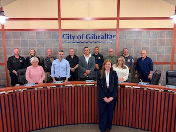 a smiling redhead in a dark suit stands in front of a long table with Gibraltar council members behind her