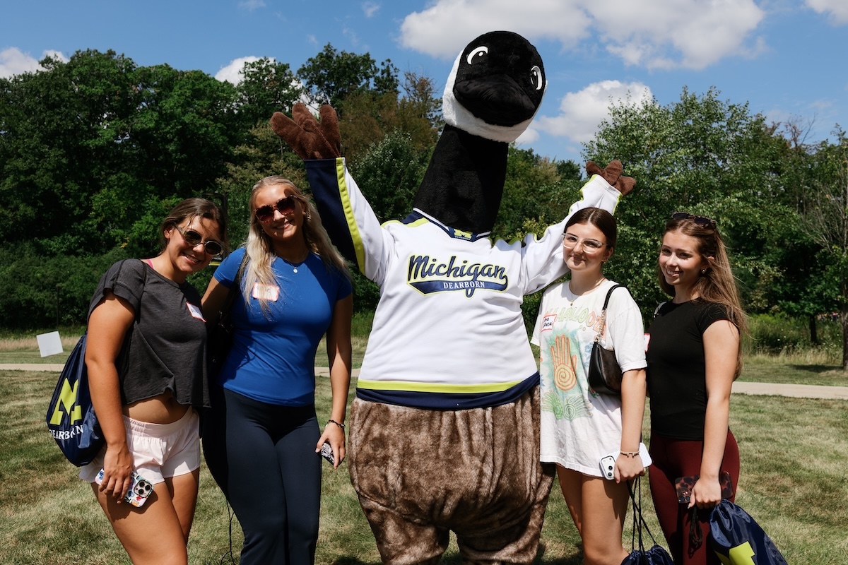 Four female students pose with the Canada goose campus friend, Bruce, outdoors on a sunny day during a campus event.