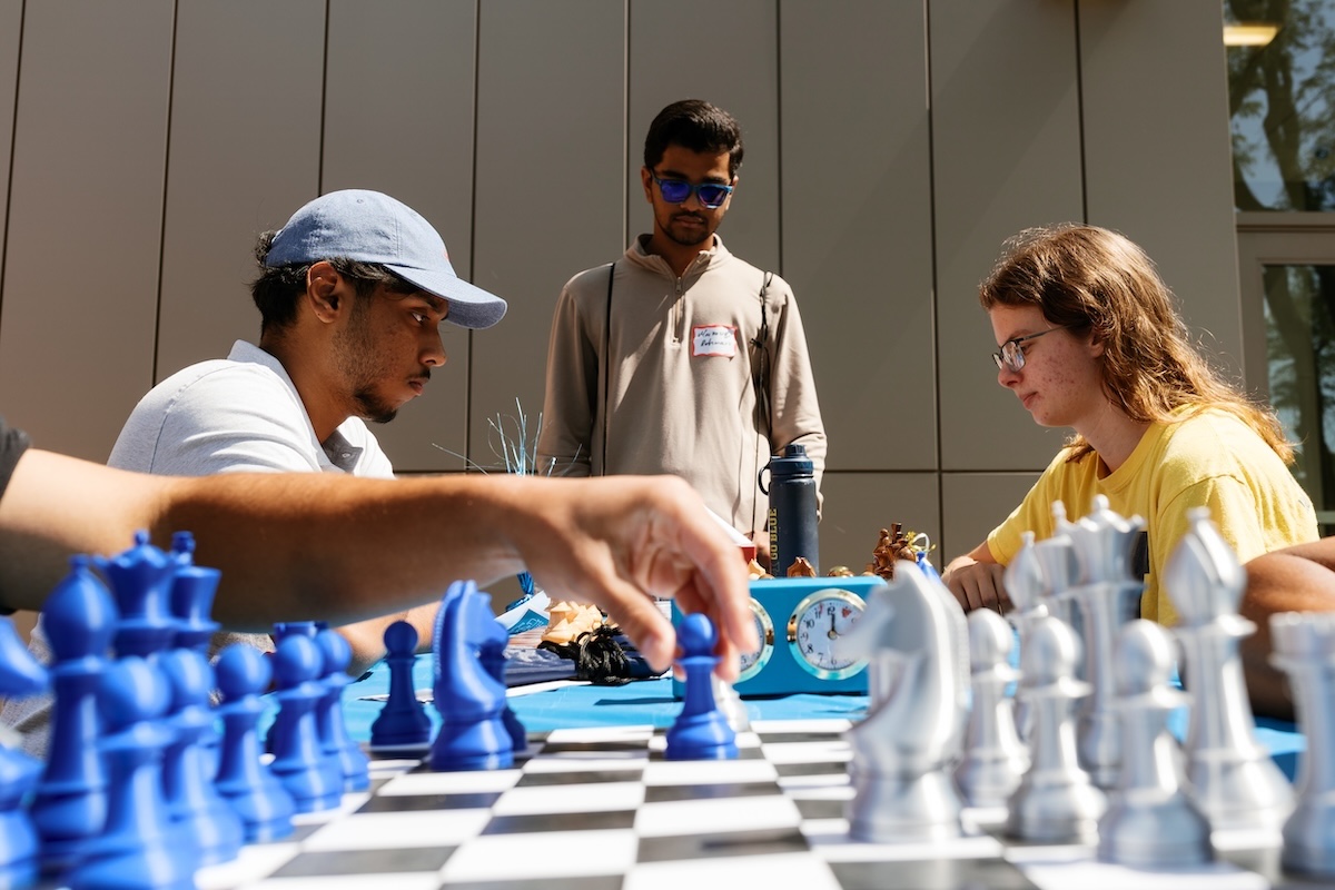 Close-up of students playing a outdoor chess game on a campus table while another student watches.