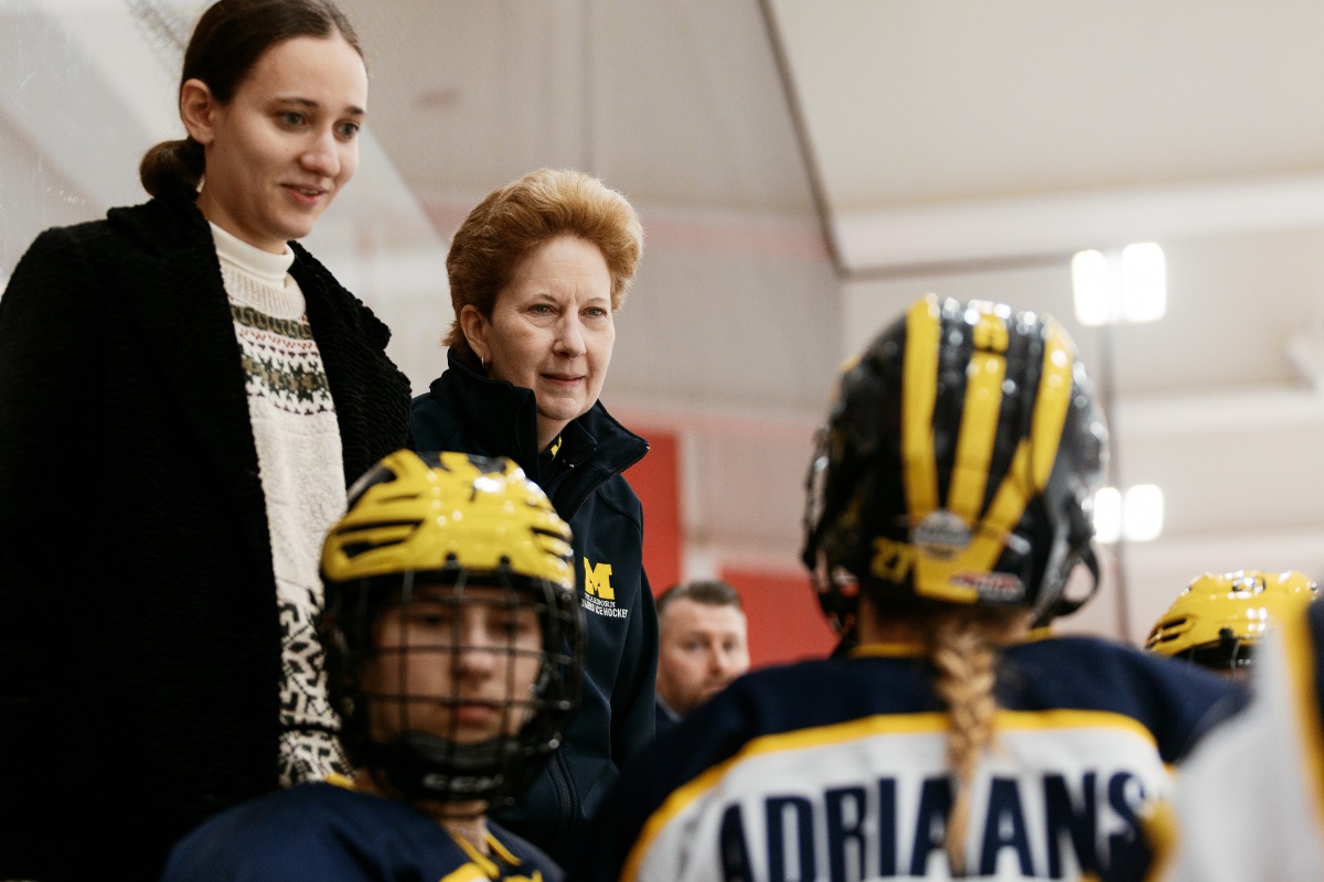 A University of Michigan-Dearborn Women’s Hockey coach and staff member stand behind players on the bench, watching the ice during a game.