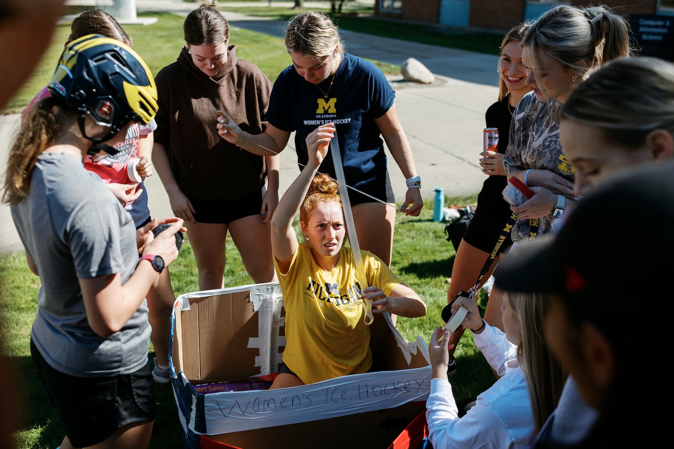 Several students gather around a cardboard boat while a woman inside the boat adds tape to keep out leaks