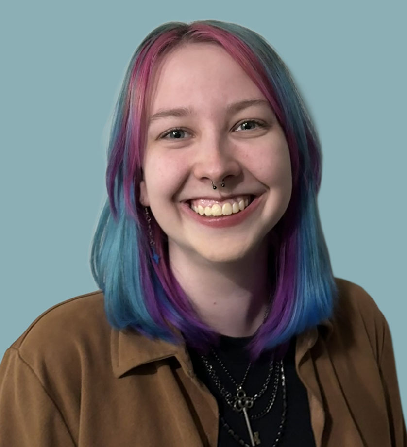 A smiling person with rainbow blue, purple and pink hair wearing a brown jacket and multiple necklaces against a blue/green backdrop