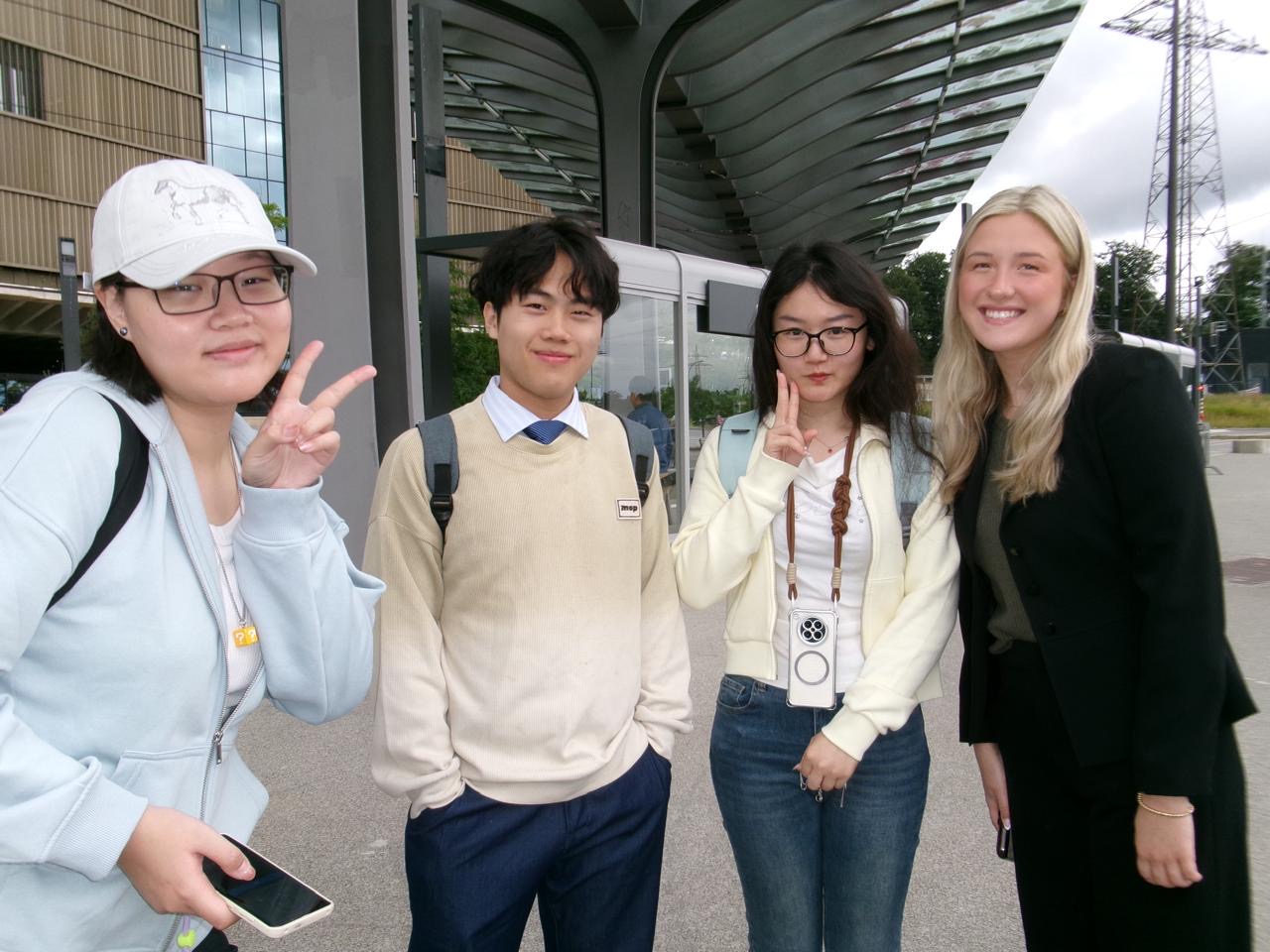 Four students pose for a photo outside a modern building with a curved metal canopy. Two students make peace signs while the group smiles at the camera.
