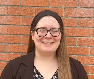 Head-and-shoulders portrait of a smiling person standing in front of a brick wall, wearing glasses, a black headband, a dark blazer, and a black floral blouse, with long hair draped over one shoulder