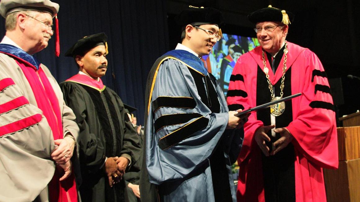 In full regalia, Xuan Zhou accepts his diploma at a commencement ceremony in front of his mentor Professor Pravansu Mohanty. 