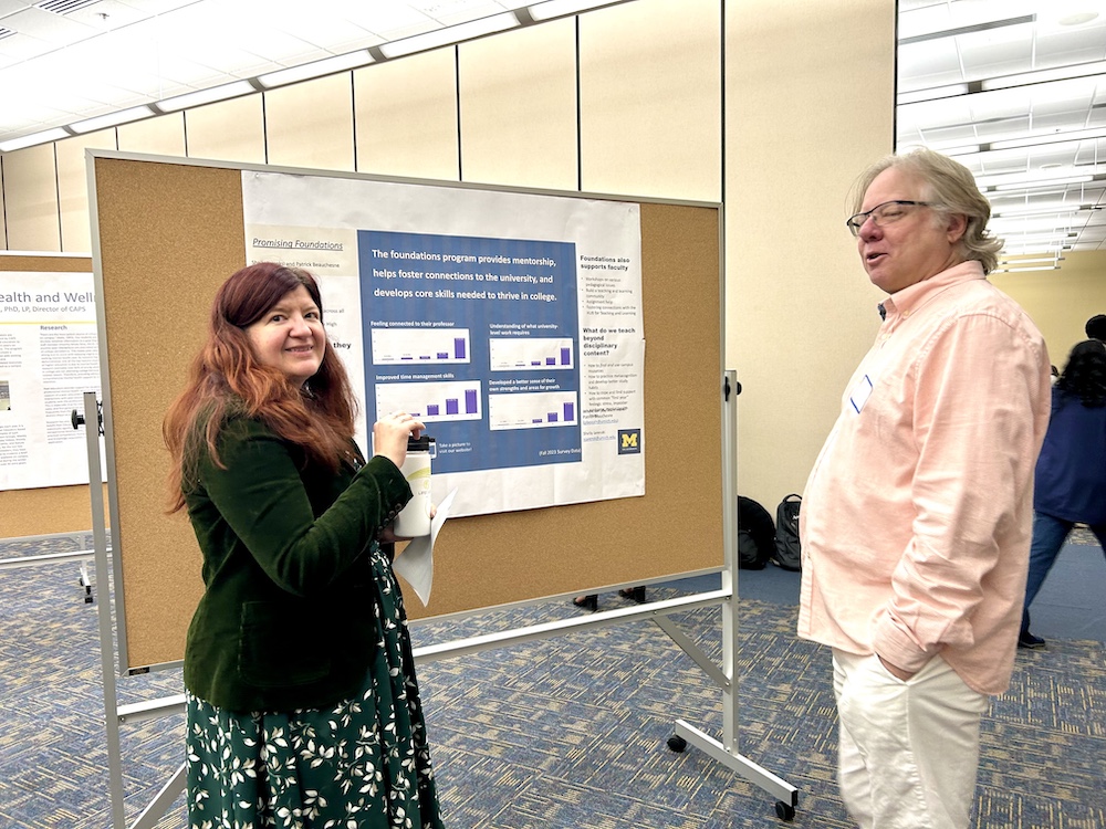 A woman with long auburn hair, dressed in a dark green jacket and green floral dress, holds a coffee tumbler while standing next to a poster board with blue and white text and bar graphs related to a university foundations program. She is in conversation with a man wearing a light pink collared shirt and light pants, who has a name tag. The setting is a large, brightly lit room with multiple academic posters displayed on cork boards on patterned carpet, suggesting a conference or research fair environment. University of Michigan branding is visible on the central poster.