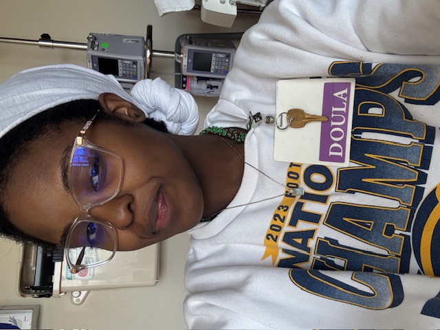 A person wearing glasses, a white headwrap, and a ‘National Champs’ sweatshirt takes a selfie indoors. They have a purple and white badge labeled ‘Doula’ clipped to their shirt. Medical equipment is visible on the wall behind them