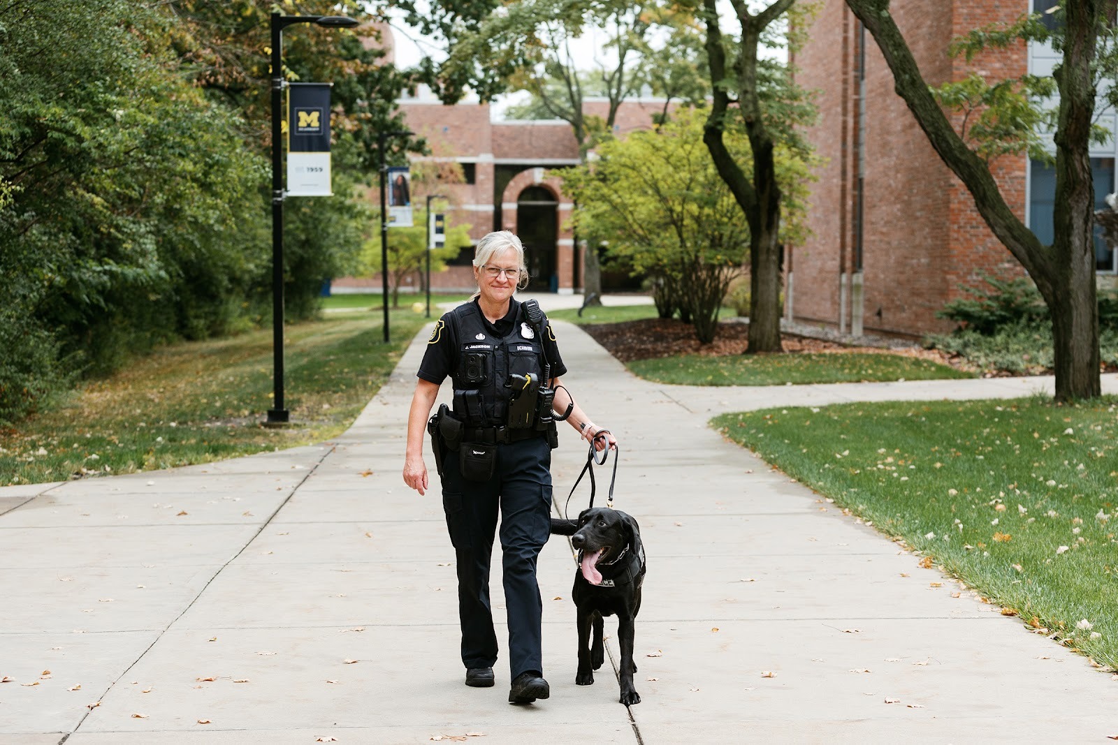 A police offficer stands next to a black lab police dog on a sidewalk on a college campus