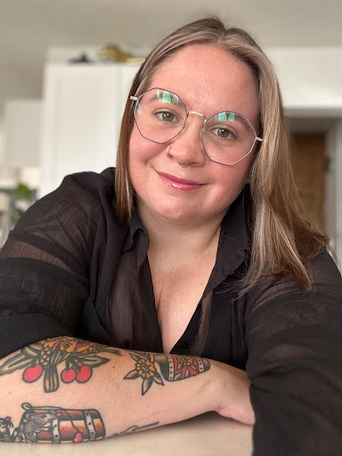 a smiling person with blondish hair wearing round glasses poses with her arms crossed in front of her in a black shirt
