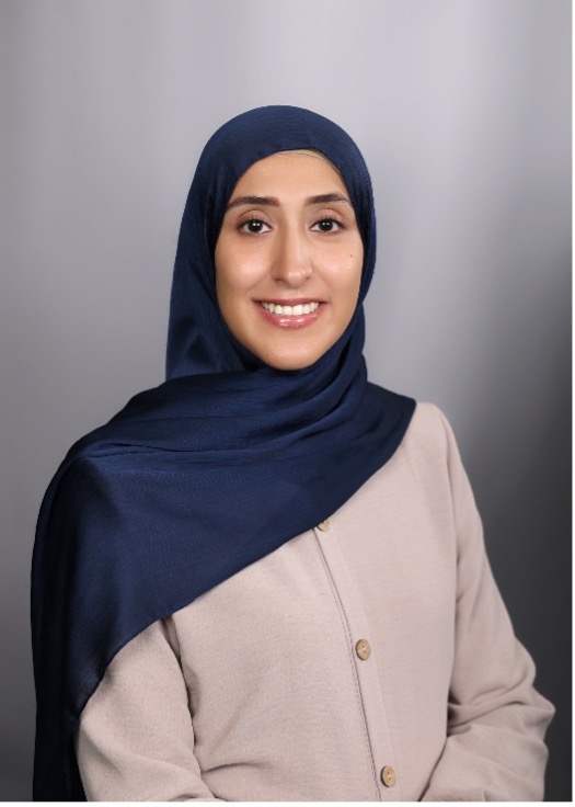 A woman smiling in a professional headshot, wearing a navy blue hijab and a light beige buttoned top, posed in front of a soft gray background.