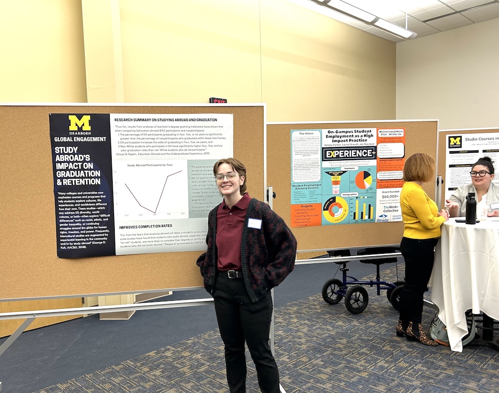 In a brightly lit academic conference room, a student wearing a red shirt and patterned cardigan, with a name tag, stands in front of a display board featuring a University of Michigan–Dearborn research poster about the impact of study abroad on graduation and retention rates. The poster includes text, a line graph, and statistics. Nearby, another board shows colorful infographics about on-campus student employment. In the background, two people converse at a high-top round table, one wearing a yellow sweater. The floor is patterned blue, and the atmosphere suggests scholarly engagement and networking.