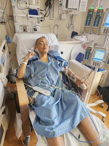 A patient in a patterned blue hospital gown rests with eyes closed in a reclined hospital chair surrounded by medical monitors and IV lines.   