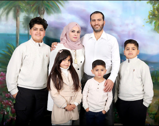 A family of six poses together in front of a painted tropical backdrop. An adult man and woman stand behind four children, with the woman wearing a pink hijab and the man in a white shirt. The children, dressed in neutral-colored outfits, stand closely together and face the camera.
