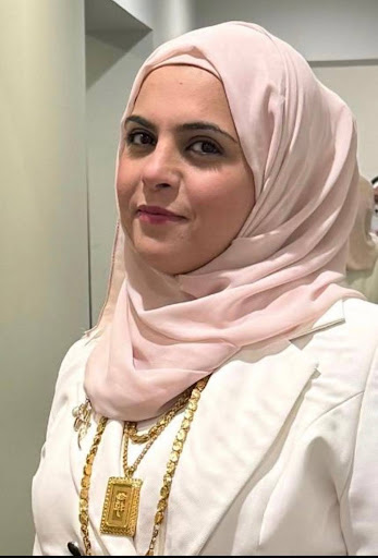A woman wearing a light pink hijab and white blazer looks toward the camera with a neutral expression. She is accessorized with layered gold necklaces and is standing indoors against a light-colored background